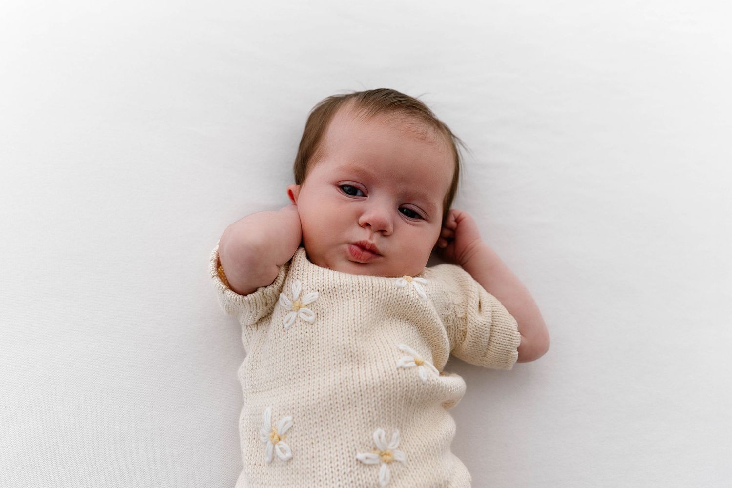 Baby in cream knit outfit touches head while lying on white background.