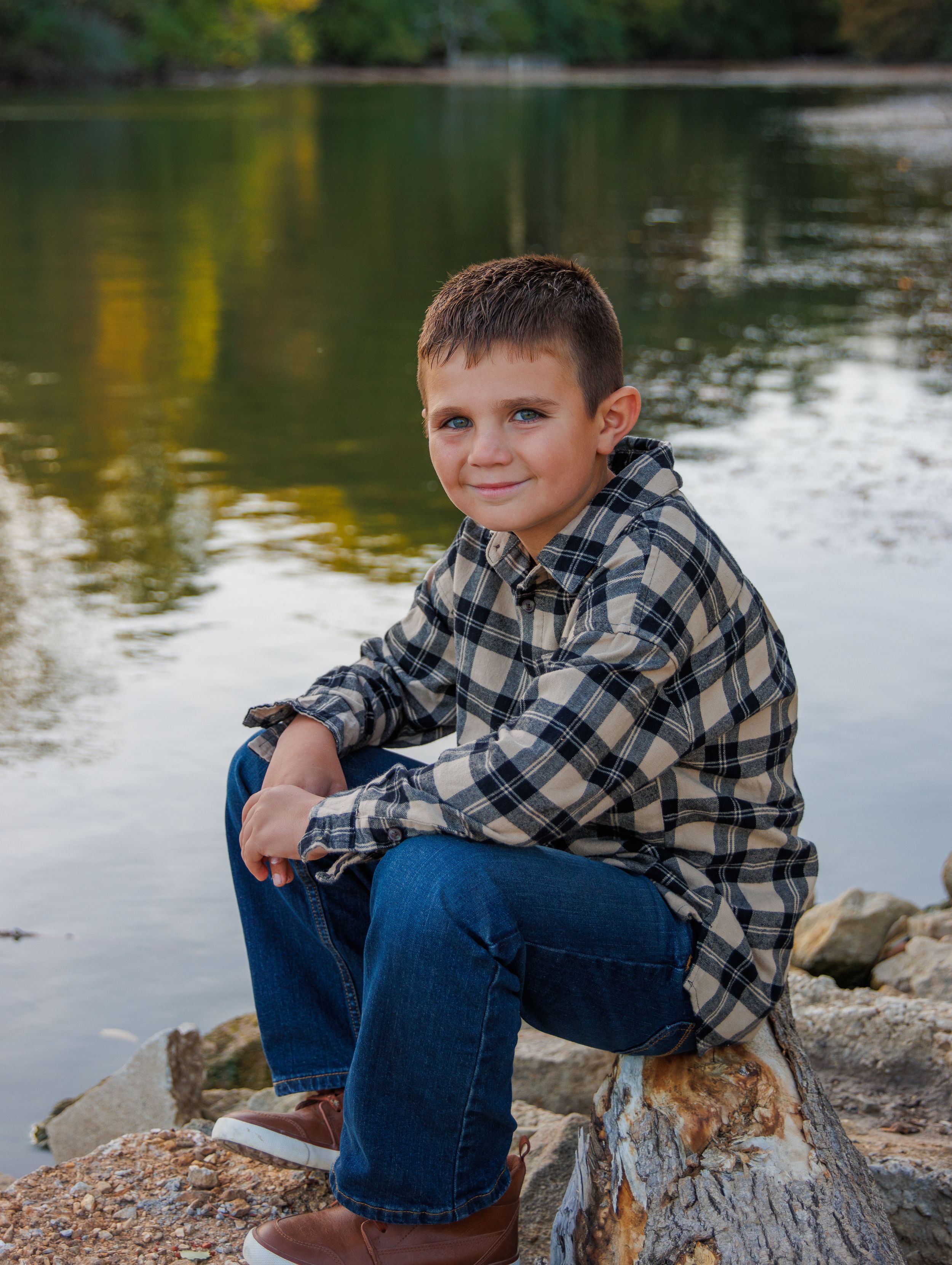 A young person in plaid sits on rocks by a serene lake during golden hour.