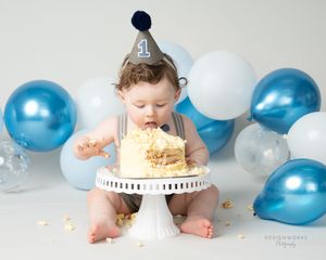 Baby in party hat smashing cake surrounded by blue and white balloons during first birthday shoot.