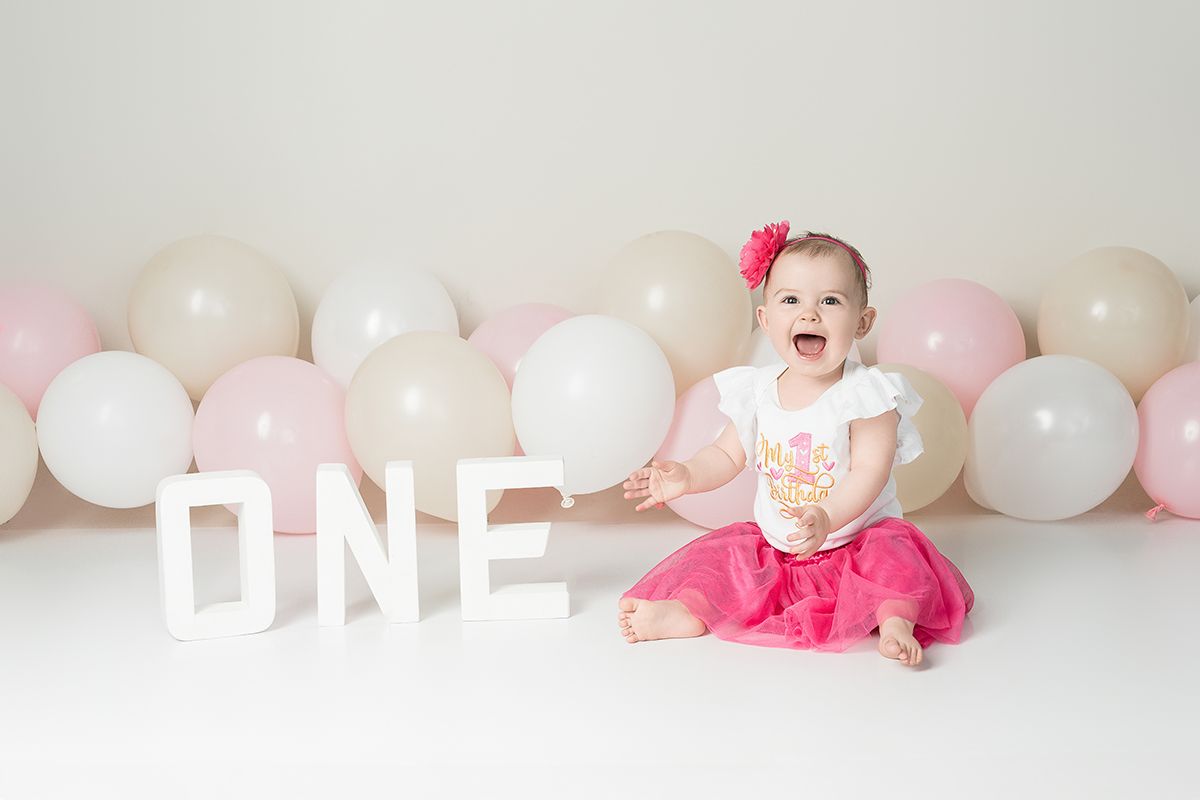 A baby wearing a pink tutu sits on the floor with white and pink balloons in the background and the word 'ONE' displayed.