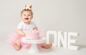 Baby in pink tutu and crown sits beside first birthday cake with white ONE letters.