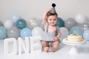 Baby in blue outfit with party hat sitting by ONE letters and birthday cake.