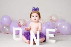 Baby in purple outfit sits with ONE letters and purple balloons for first birthday photo.