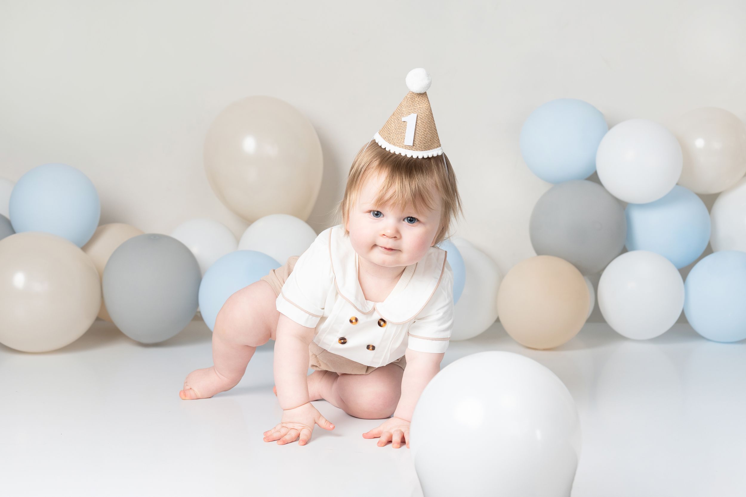 Baby in party hat crawling among blue and gray balloons during first birthday photo shoot.