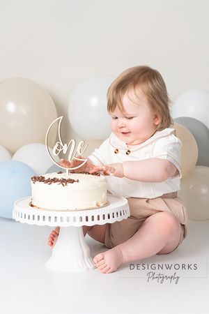 Baby reaching for white birthday cake on pedestal with balloon backdrop.