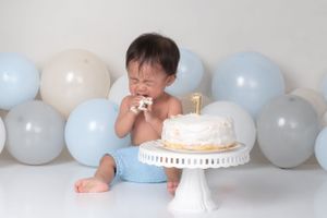 Baby in blue outfit with birthday cake on white pedestal and balloon background.