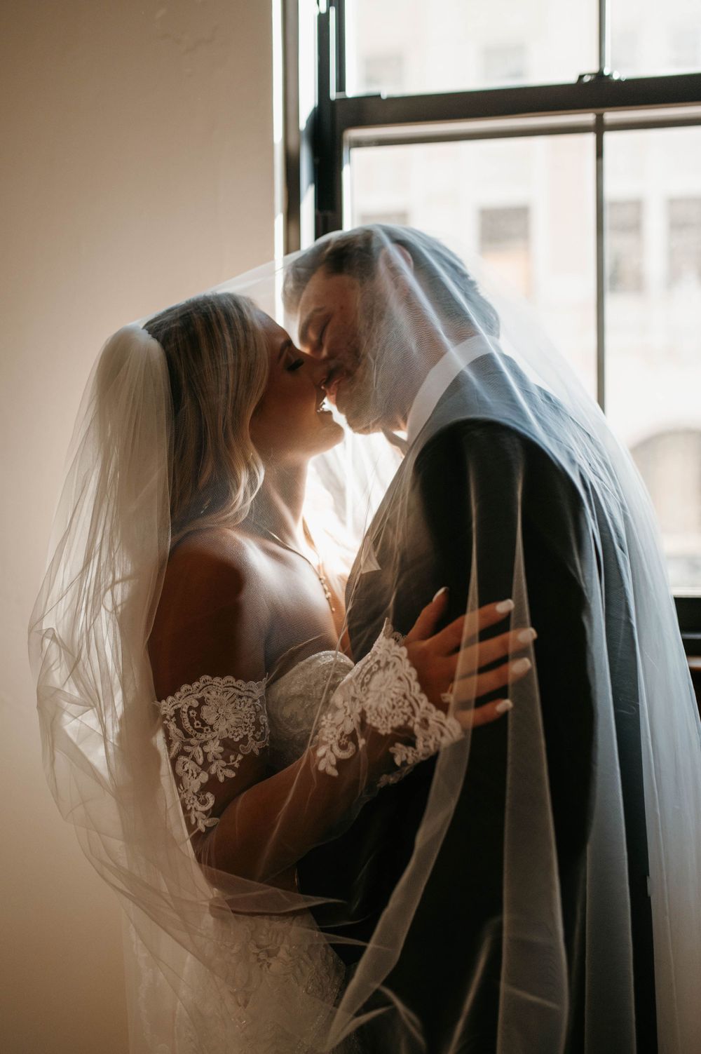 Romantic sunlit wedding scene with a bride and groom sharing an intimate moment by a window in elegant attire. The Treasury