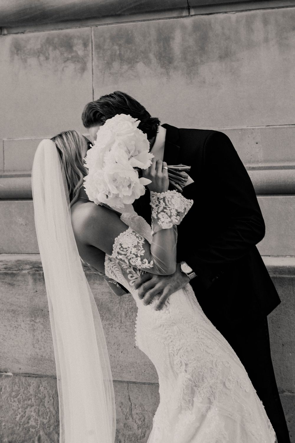 A romantic black and white wedding photograph showing a couple kissing behind a white peony bouquet.