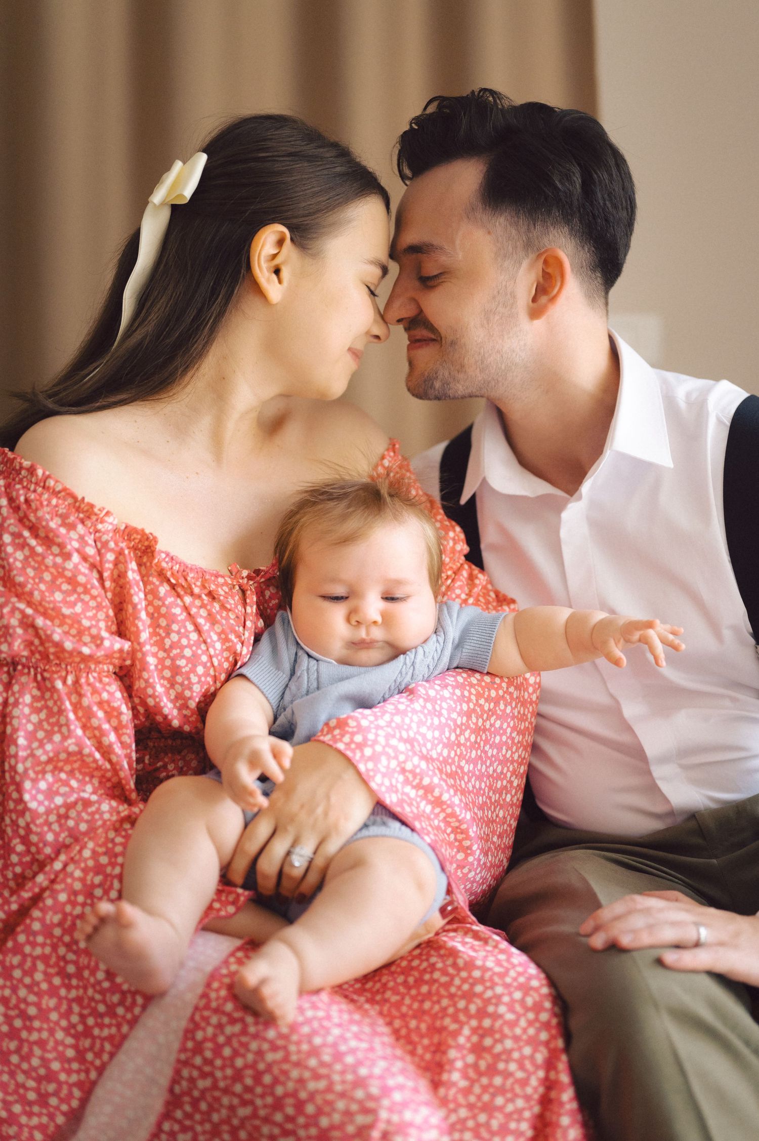 Intimate family moment with parents lovingly embracing while holding their smiling baby in a warm interior setting.