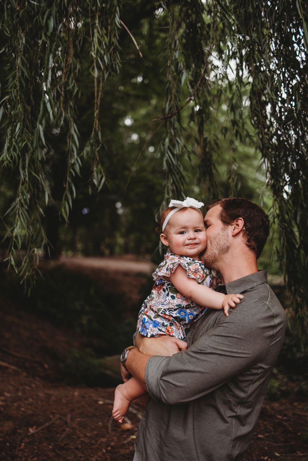 A parent holding and kissing a baby under weeping willow trees.
