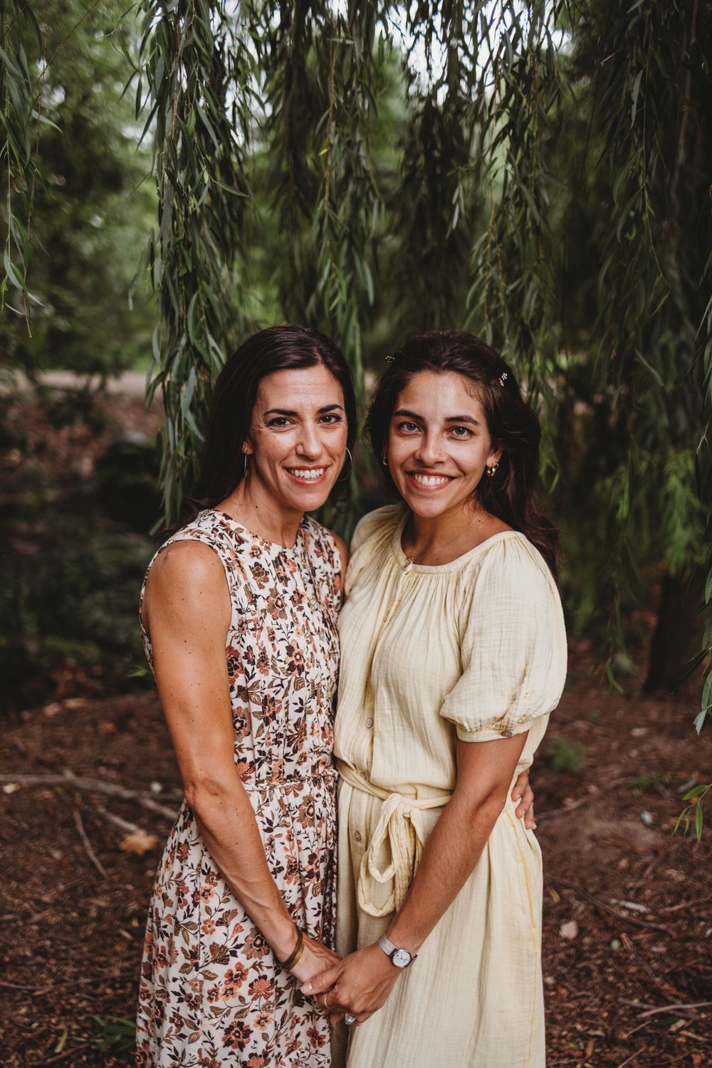 Two women in summer dresses pose together under a weeping willow tree.