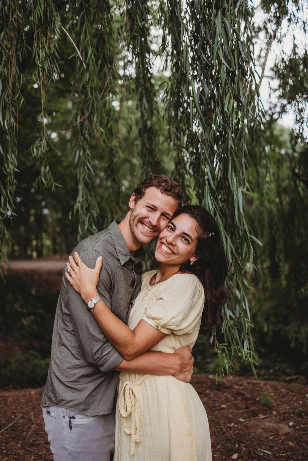 A couple embracing and smiling under weeping willow trees.