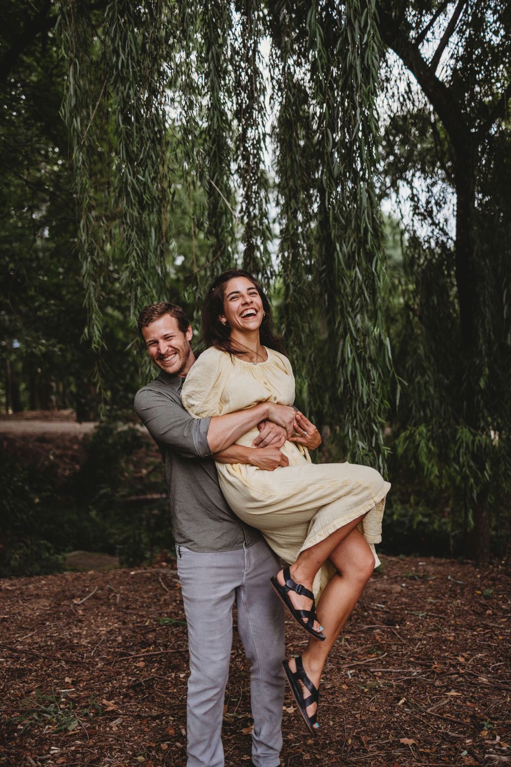 Playful couple laughs while sharing a candid moment under willow tree branches.