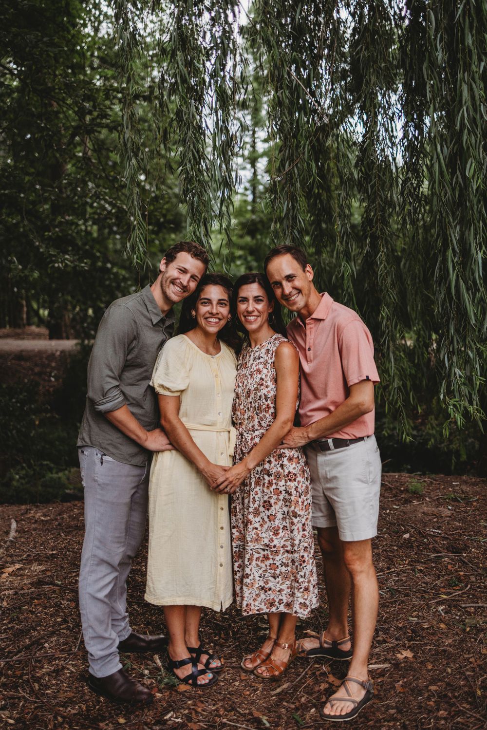 Group portrait of four people smiling beneath willow trees on a wooded path.