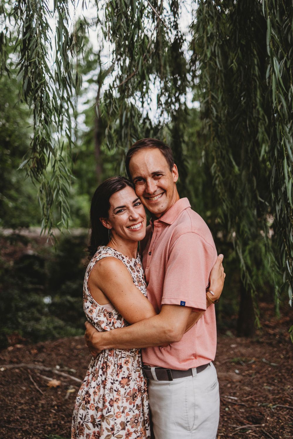 Couple embracing and smiling in casual attire under willow tree branches.