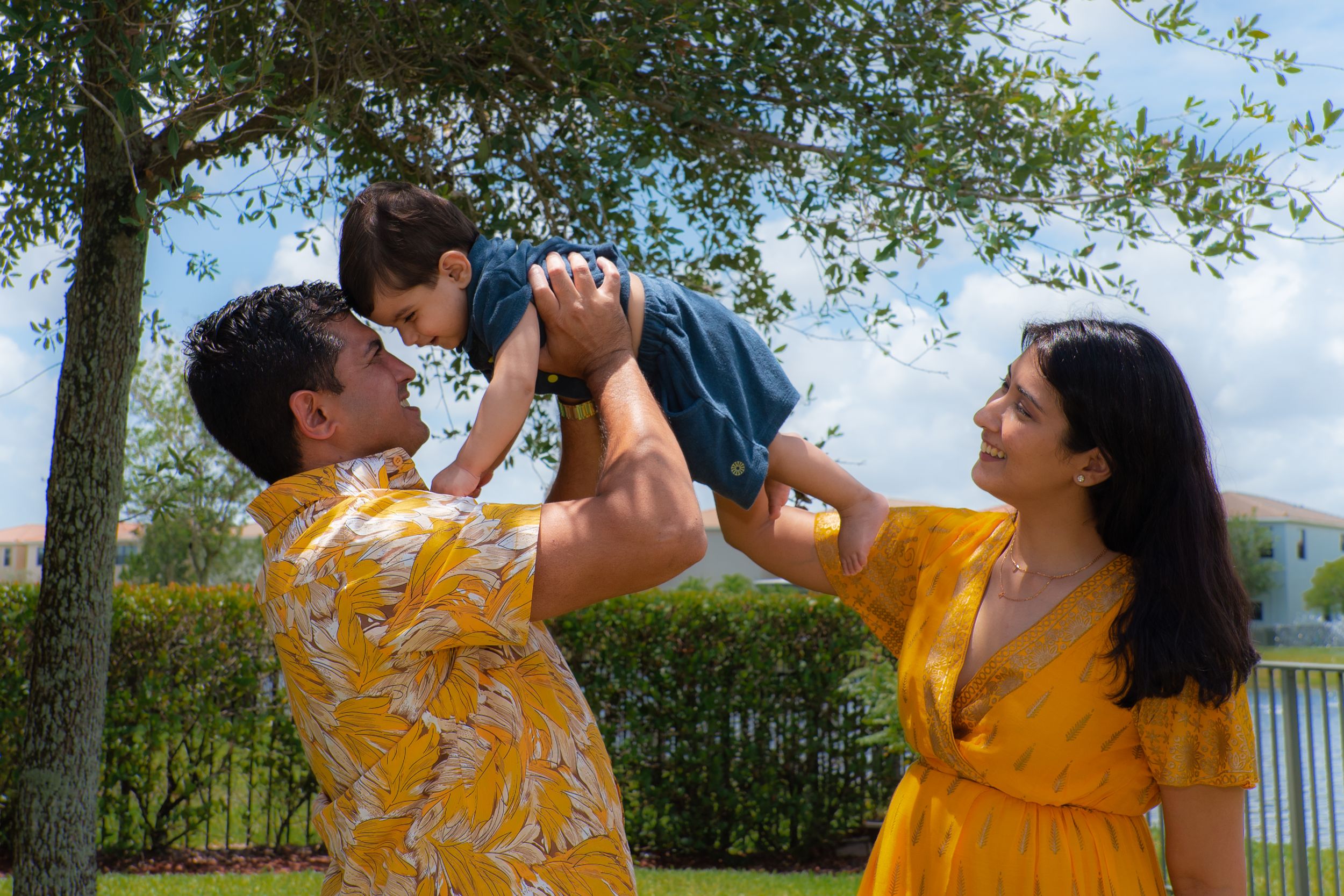 Parents joyfully lift their toddler son outdoors near a tree in matching yellow outfits.