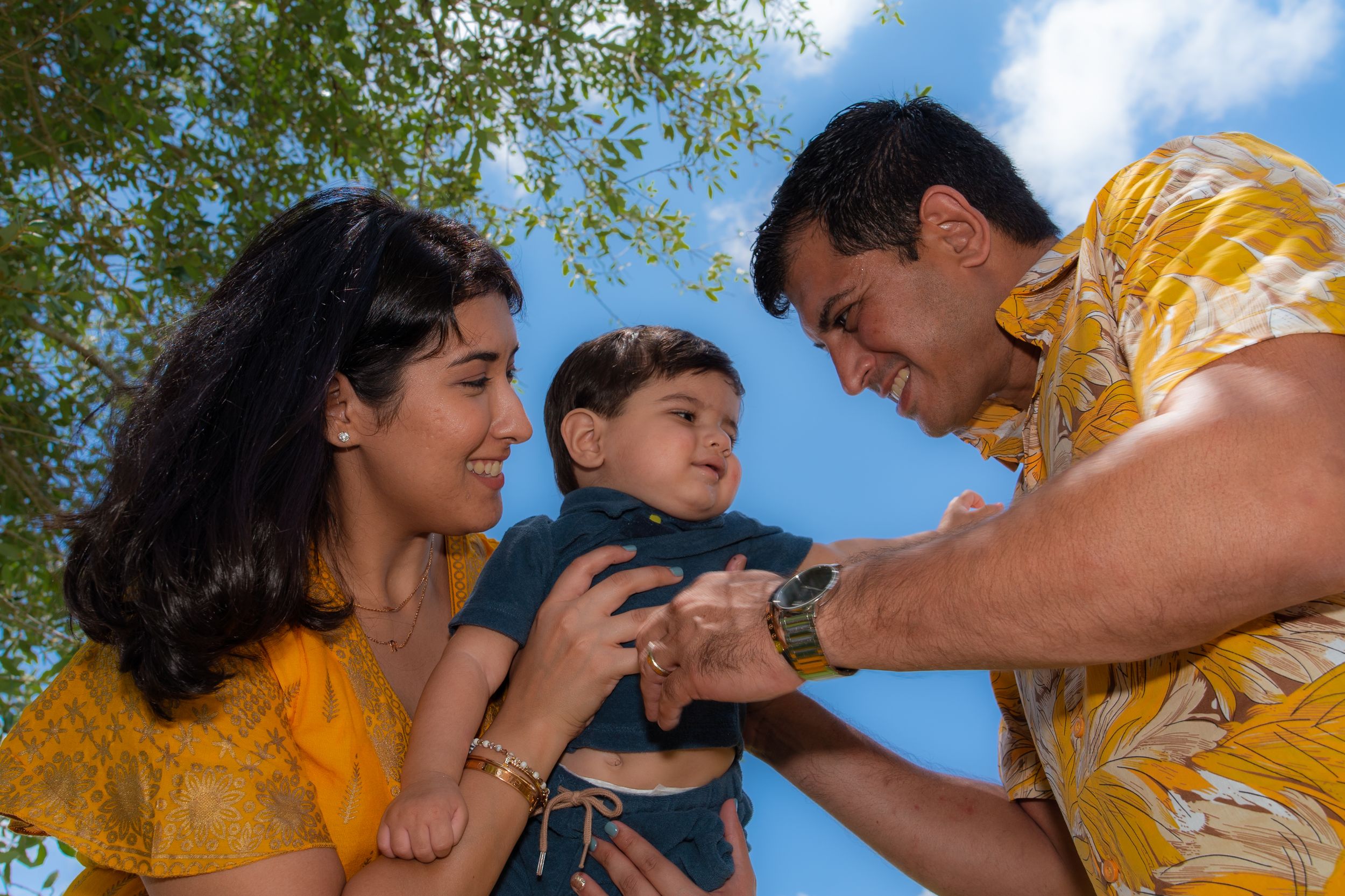 Happy couple shares a tender moment with their smiling baby boy under a blue sky.