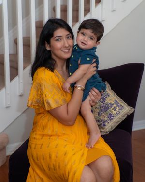 Woman in yellow dress holding baby boy in teal outfit while seated in purple chair.