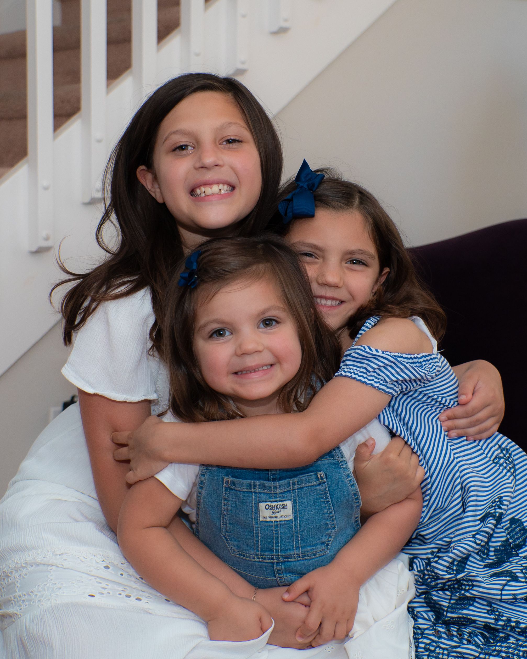 Three smiling sisters pose together on stairs, wearing blue hair bows and coordinating outfits.