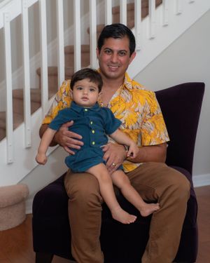 Smiling man in yellow floral shirt sitting in purple chair holding baby boy indoors.