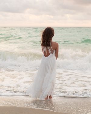 Woman in white dress standing at the ocean's edge viewed from behind.