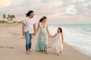Family maternity beach photoshoot with pregnant woman in mint dress, man, and young girl walking along sandy shore.