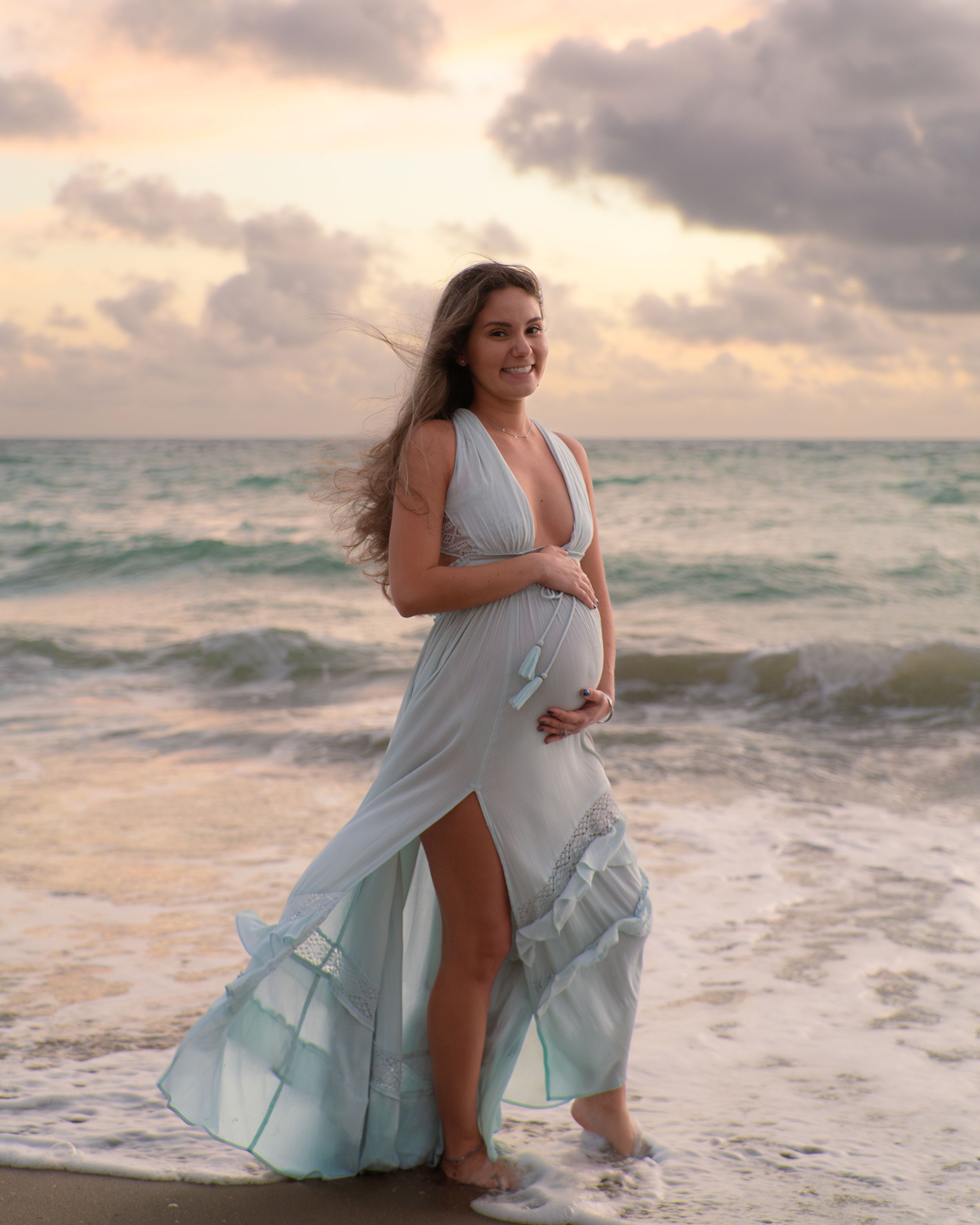 Pregnant woman in light blue gown posing on beach at sunset.
