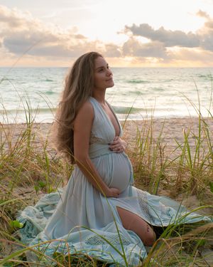 Pregnant woman in flowing mint dress sitting in beach grass at sunset during maternity photoshoot.