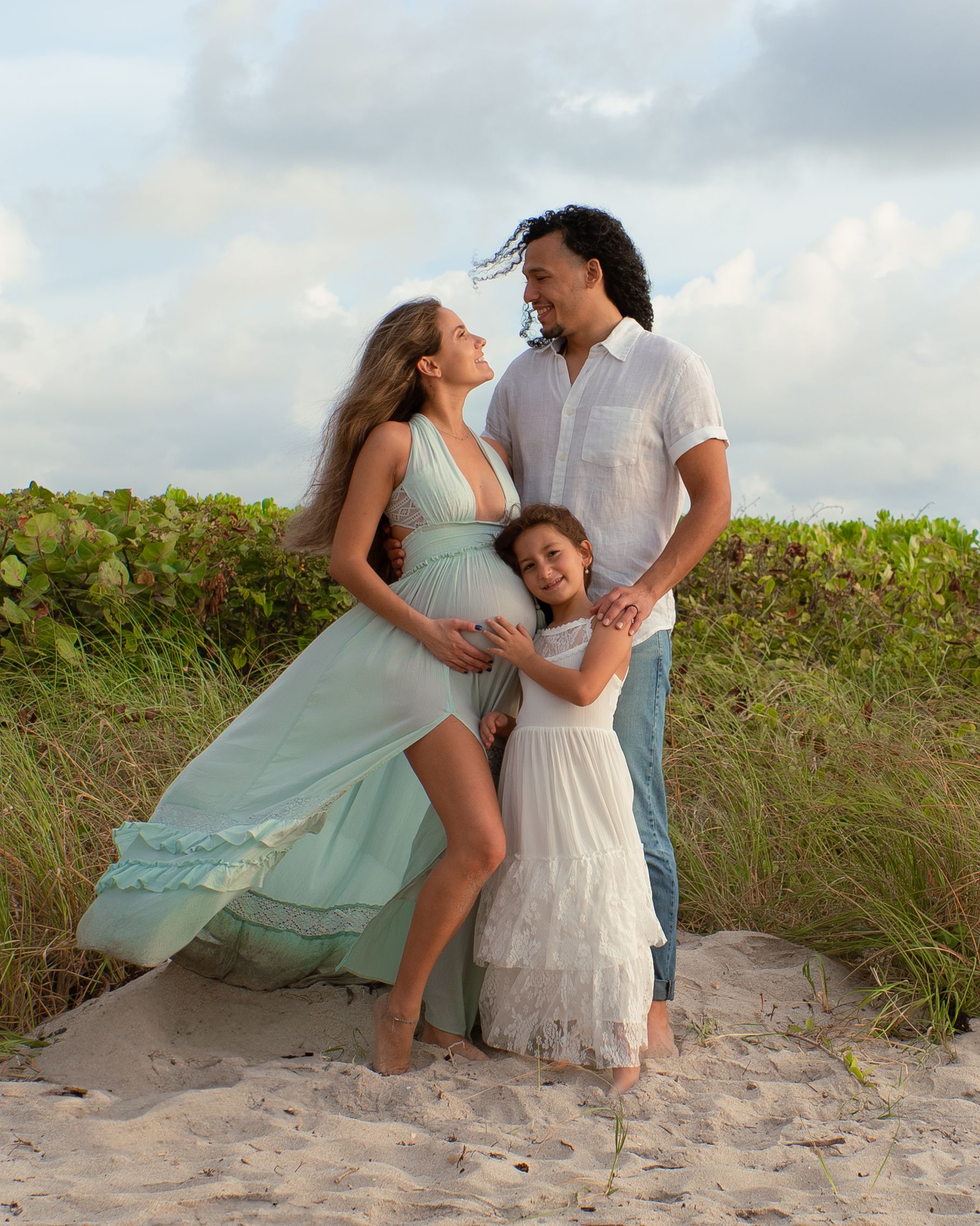 Family maternity photo with pregnant woman in mint dress, long-haired man, and young girl on beach.