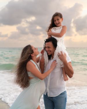 Family laughing on beach with father lifting daughter on his shoulders.