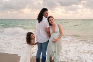 Pregnant couple with daughter posing together on a scenic beach.