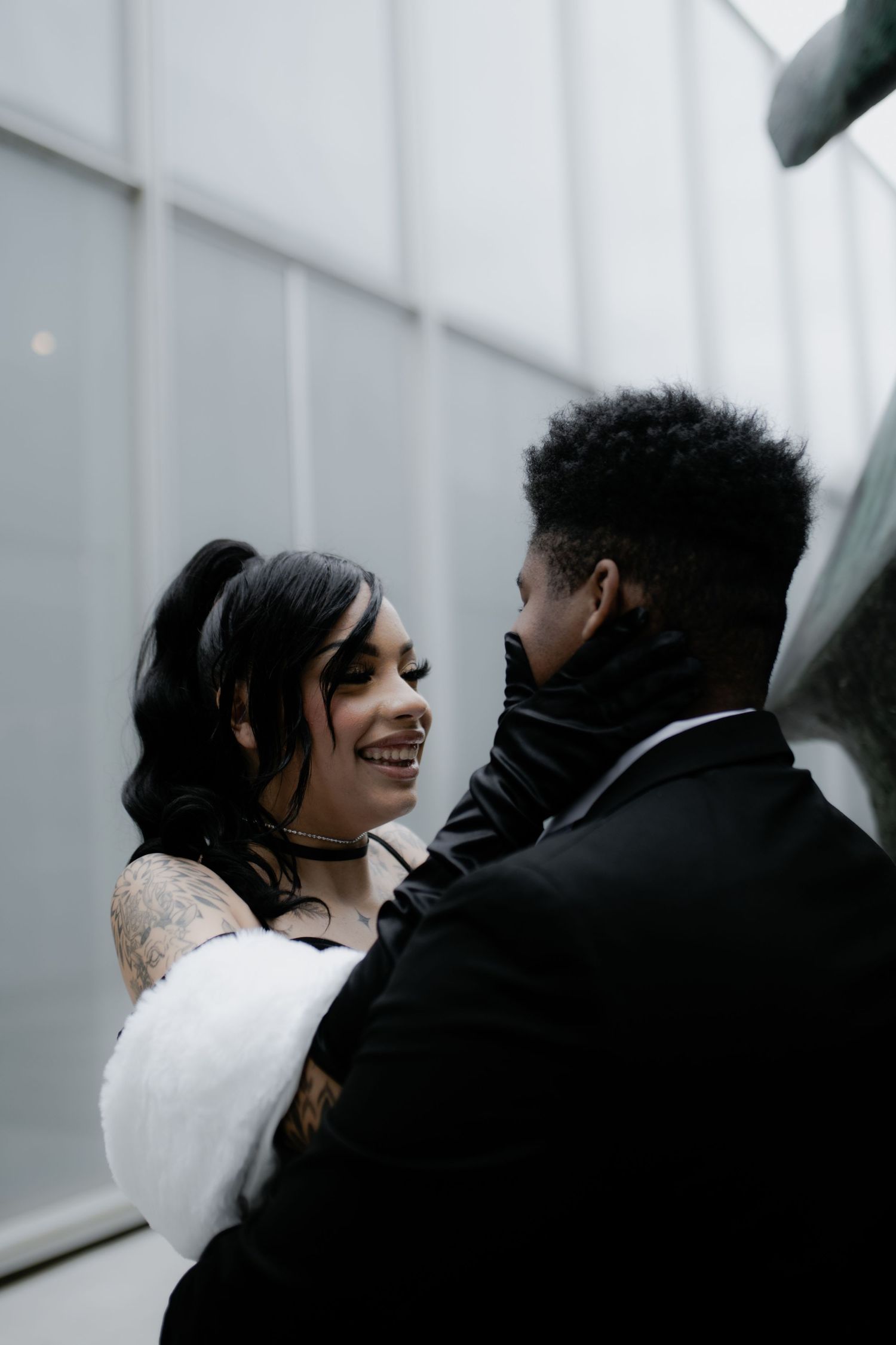 Couple in formal attire embracing against minimalist white architecture.