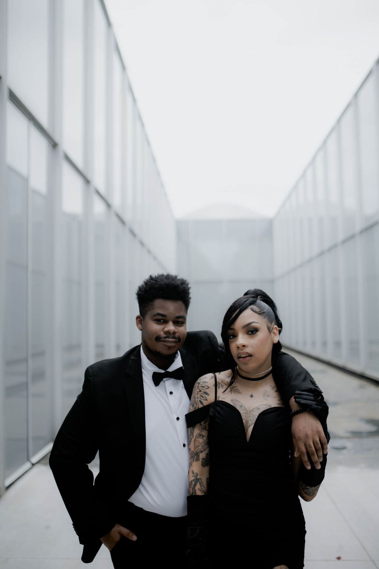 Couple in black formal wear posing against modern white architectural backdrop.