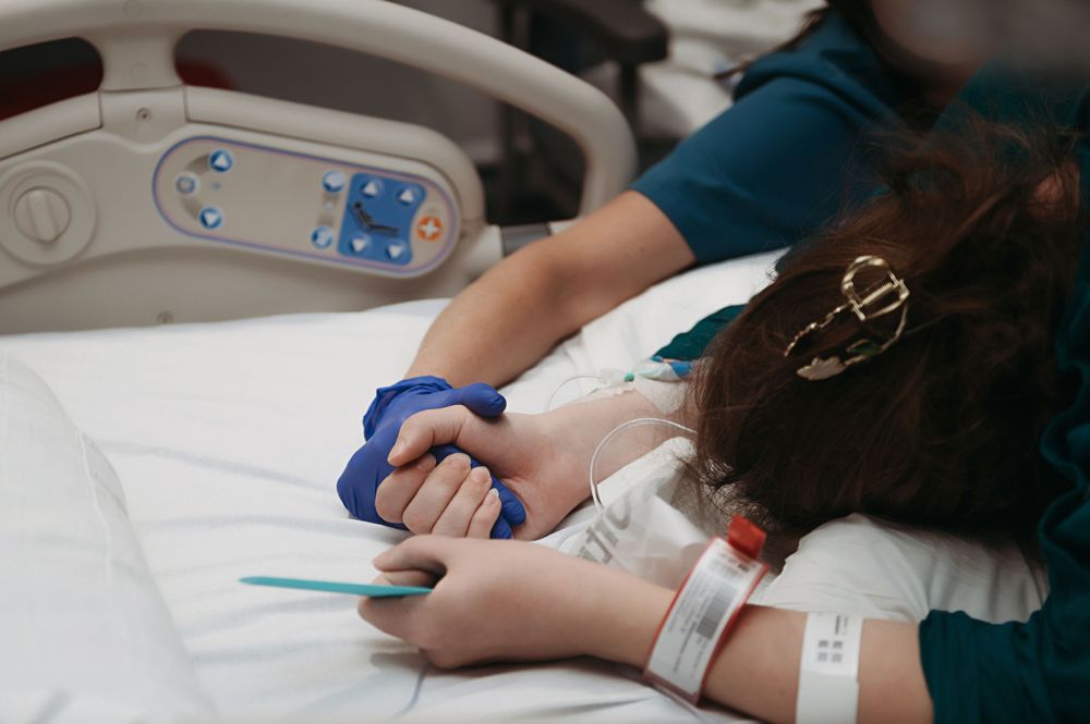 Someone holds hands with a patient in a hospital bed with medical equipment and monitors visible.