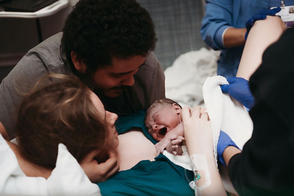 A newborn baby cries while being comforted by family members and medical staff in a hospital setting.