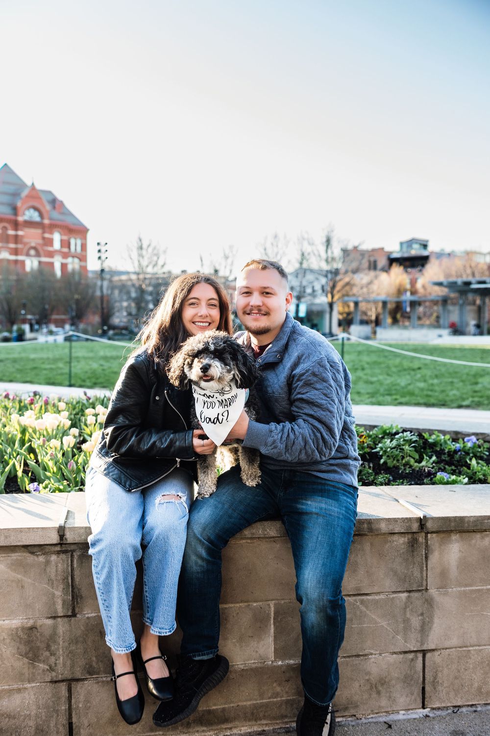Happy engaged couple sitting on stone wall holding fluffy dog with proposal bandana outdoors.