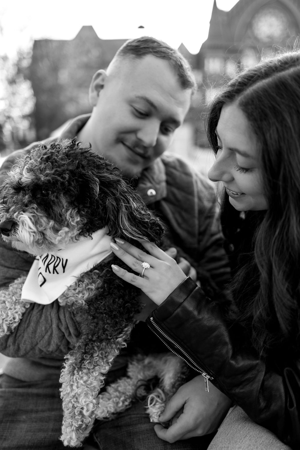 Black and white photo of smiling couple cuddling fluffy dog with proposal bandana outdoors.