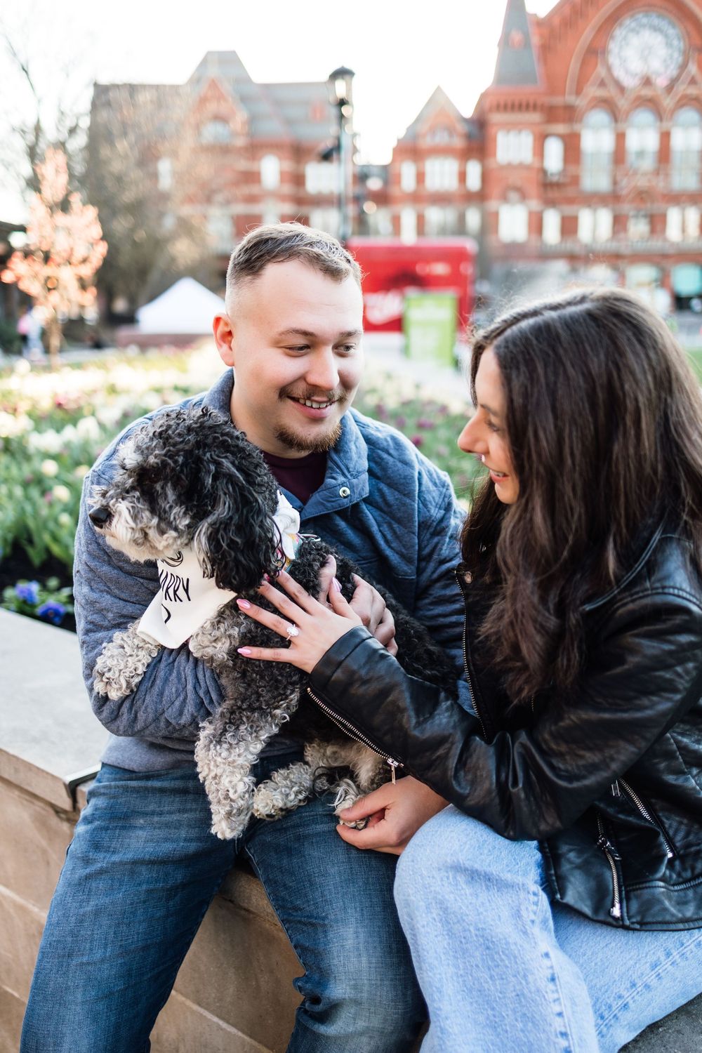 Couple smiling at their small fluffy dog in an urban park setting with brick building background.