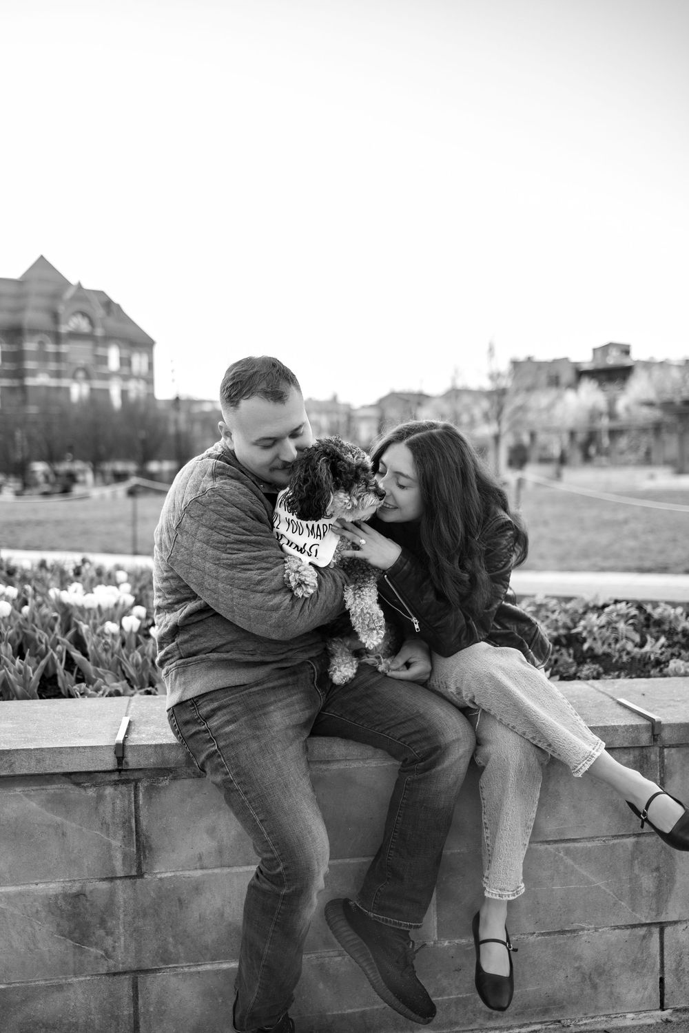 Black and white photo of couple sitting on stone wall sharing kiss with small fluffy dog.