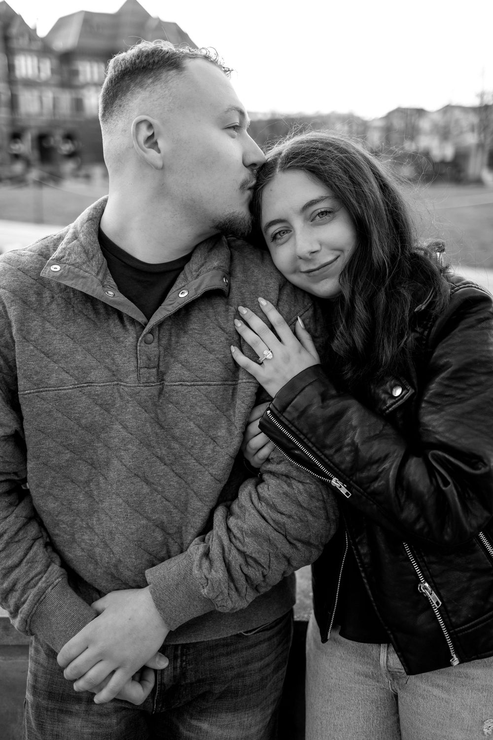Black and white photo of man kissing woman's temple while she shows her engagement ring.