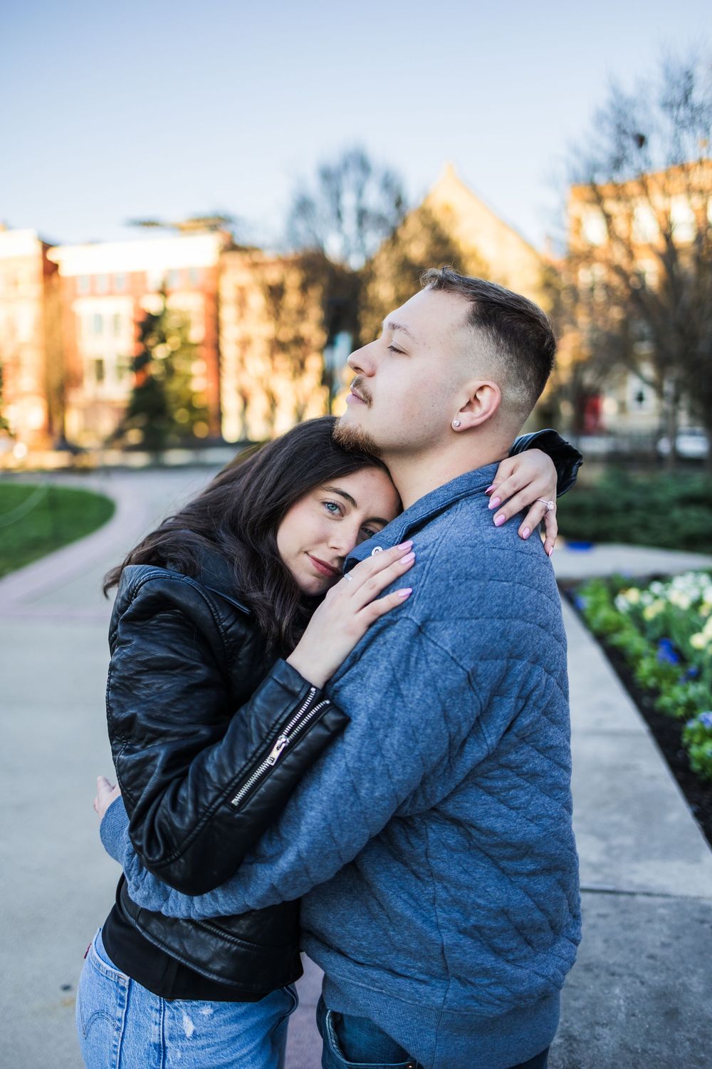 Happy couple embracing tightly outdoors in a park with buildings and flowers in background.