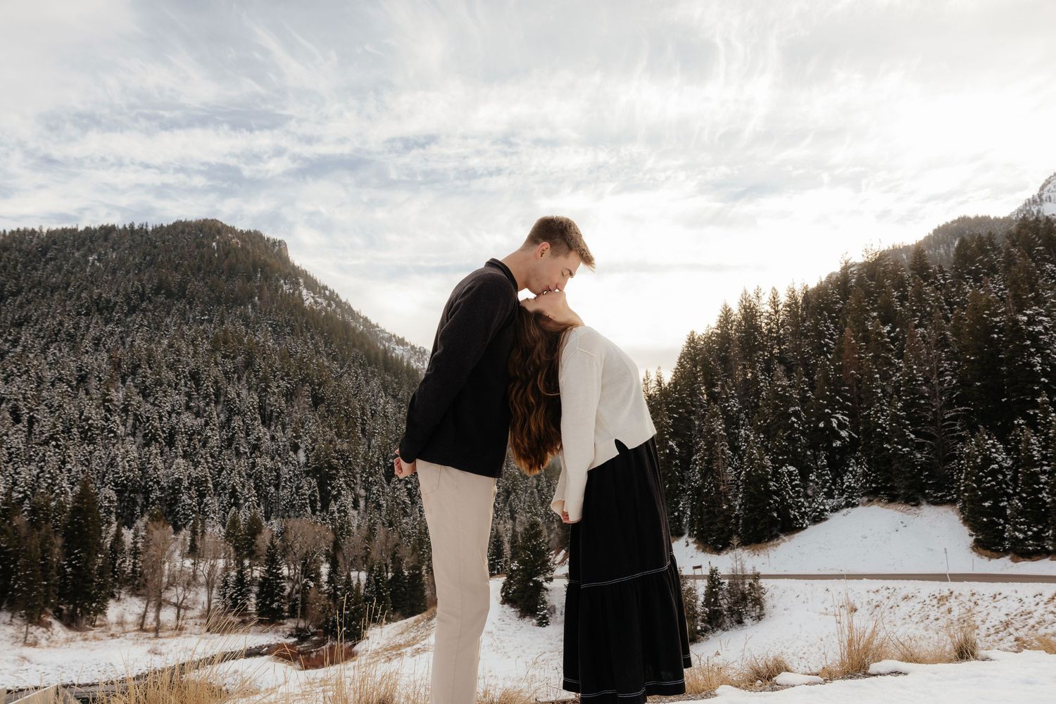 Couple holding hands and touching foreheads in snowy mountain landscape with pine trees.
