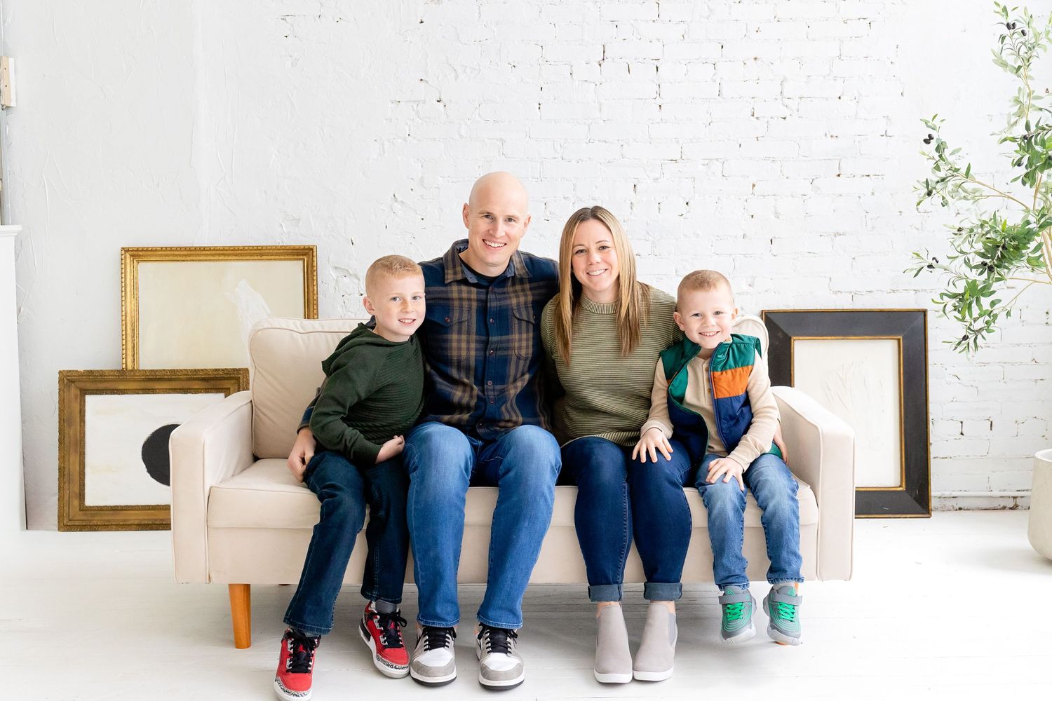A happy family of four sits together on a white couch with empty picture frames in the background.