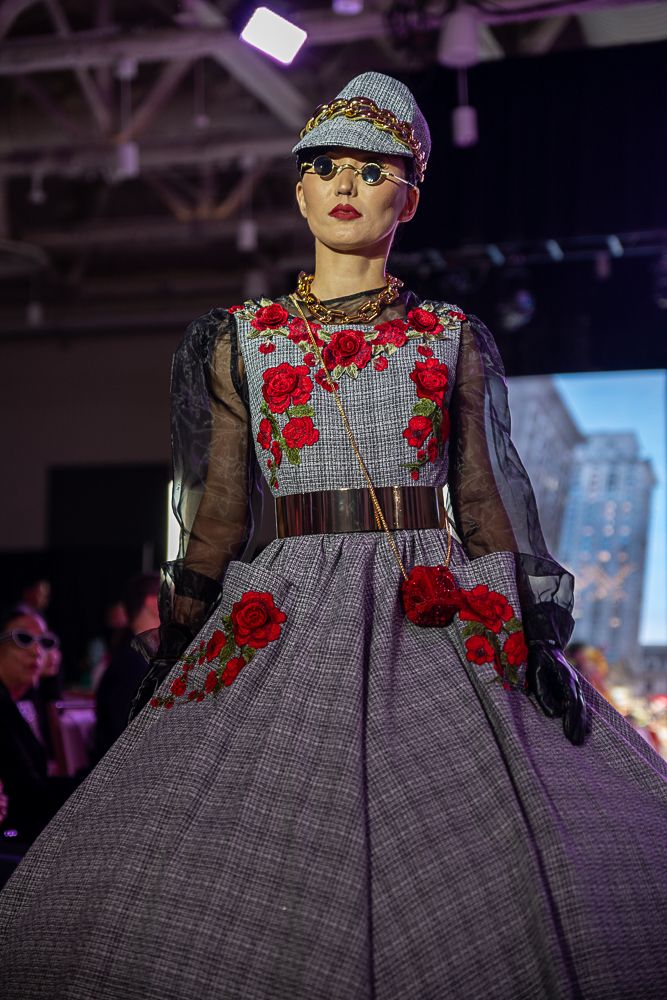 Fashion model on runway wearing gray dress with red floral appliques, sheer sleeves and metallic cap accessory.