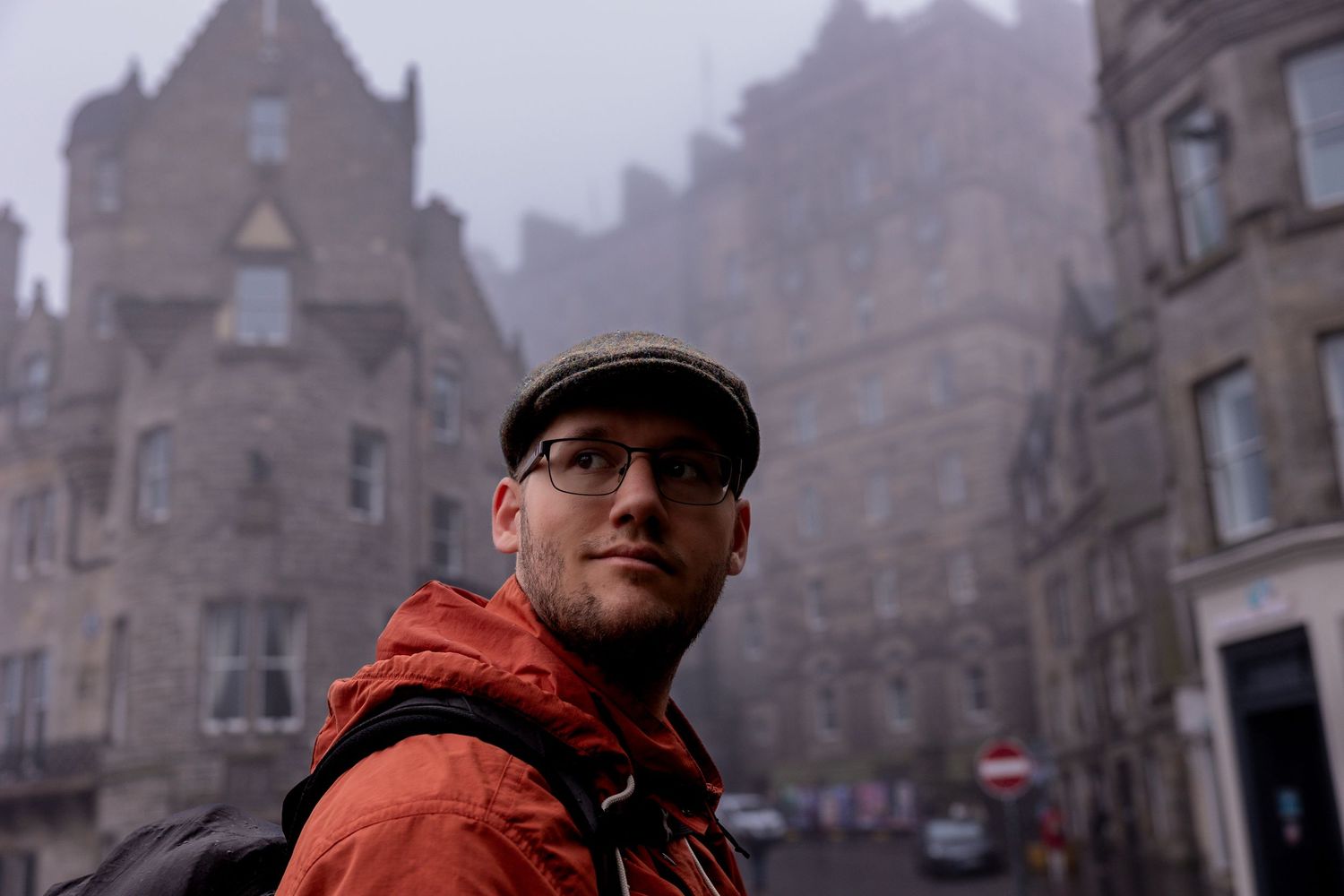 Person in orange jacket and flat cap stands against misty backdrop of historic Edinburgh architecture.
