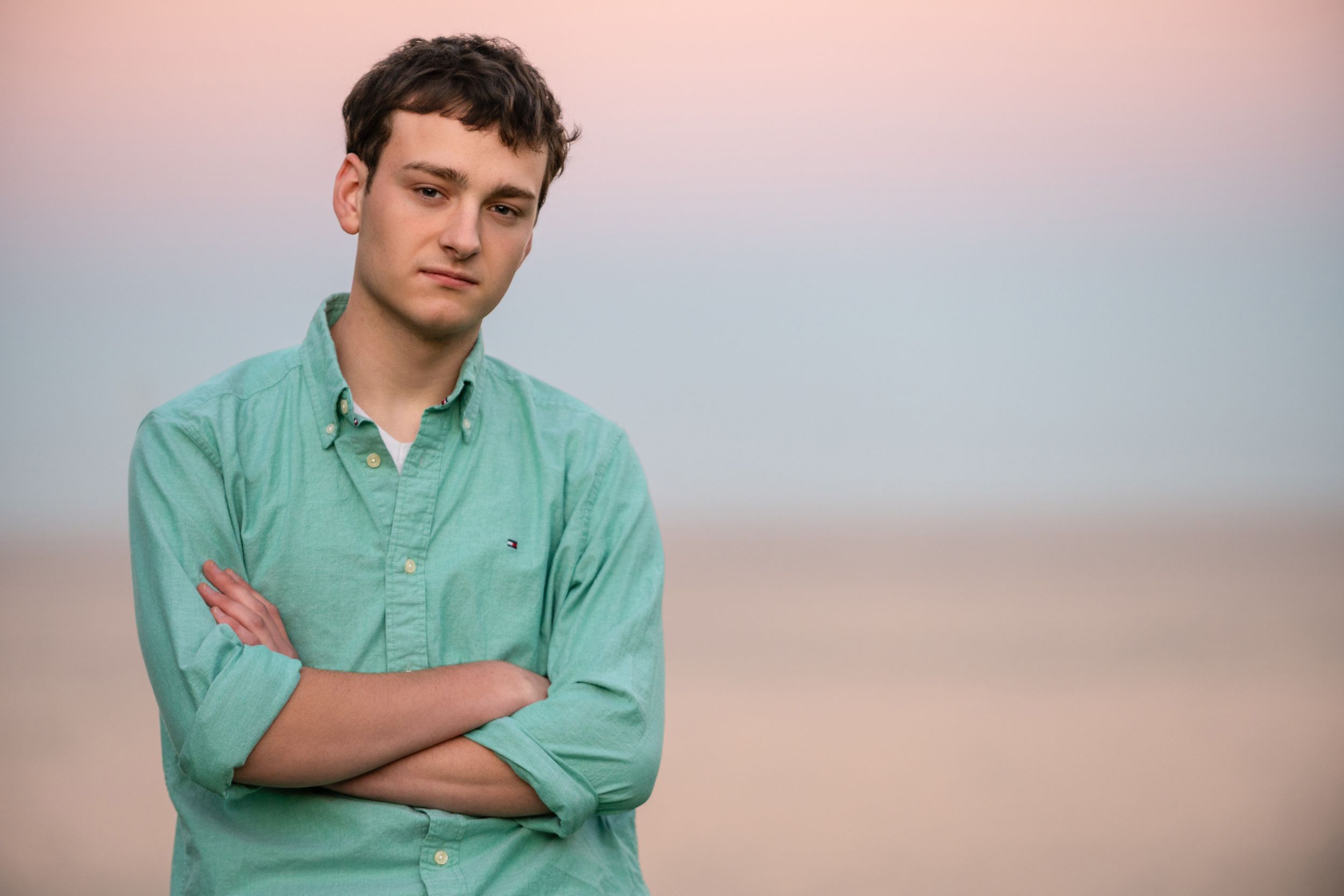 Young man in teal shirt standing with arms crossed on a beach at sunset.
