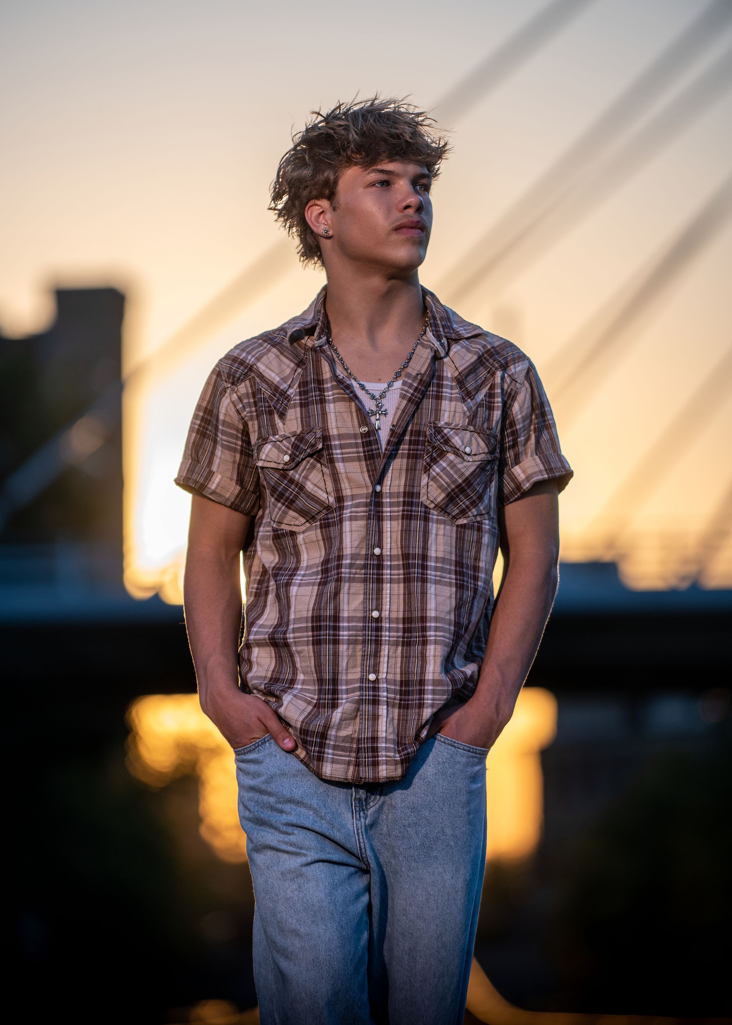Young man in plaid shirt and jeans poses outdoors at golden hour with a bridge silhouette in the background.