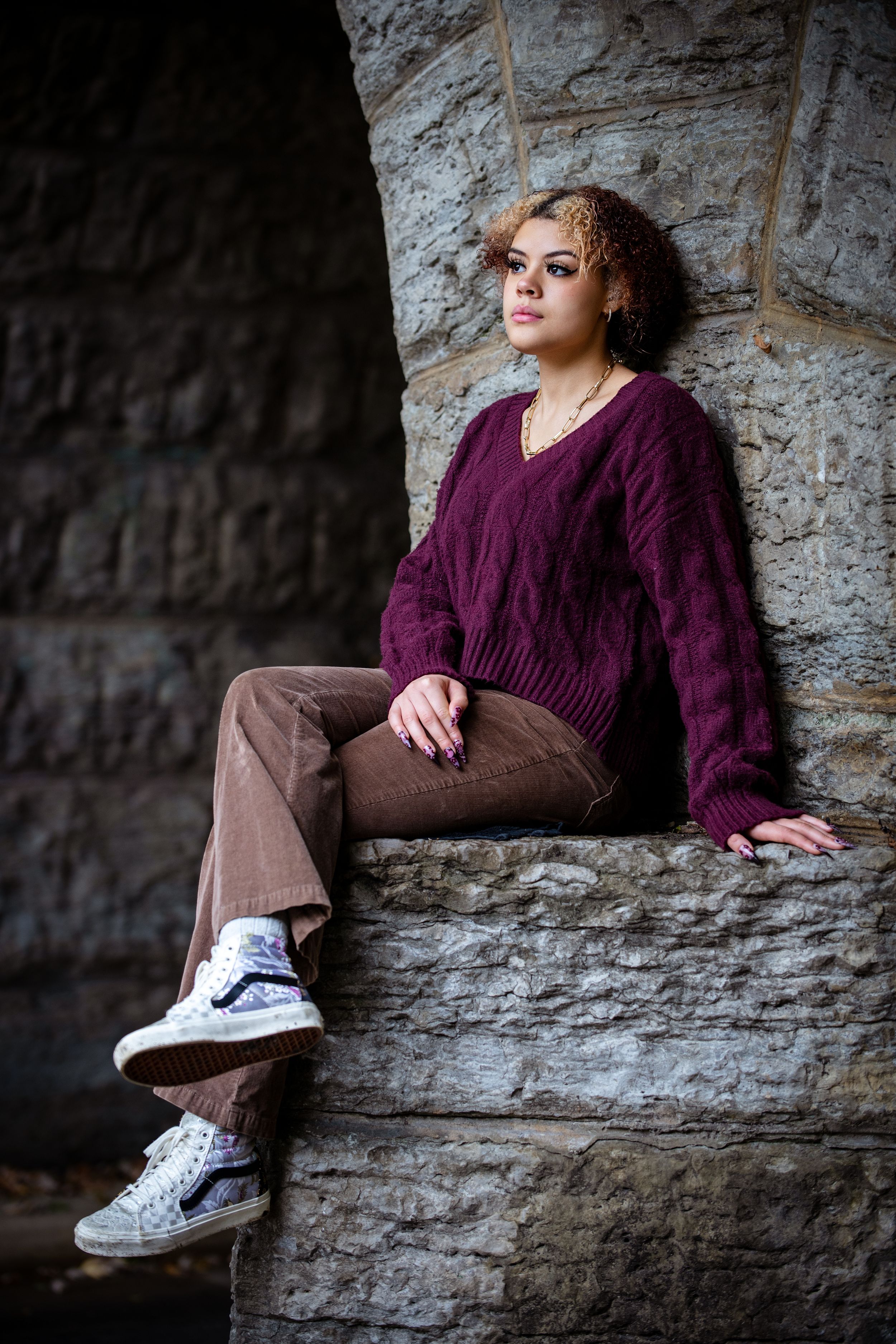 Young woman in burgundy cable-knit sweater and brown pants sits on stone wall wearing white sneakers.