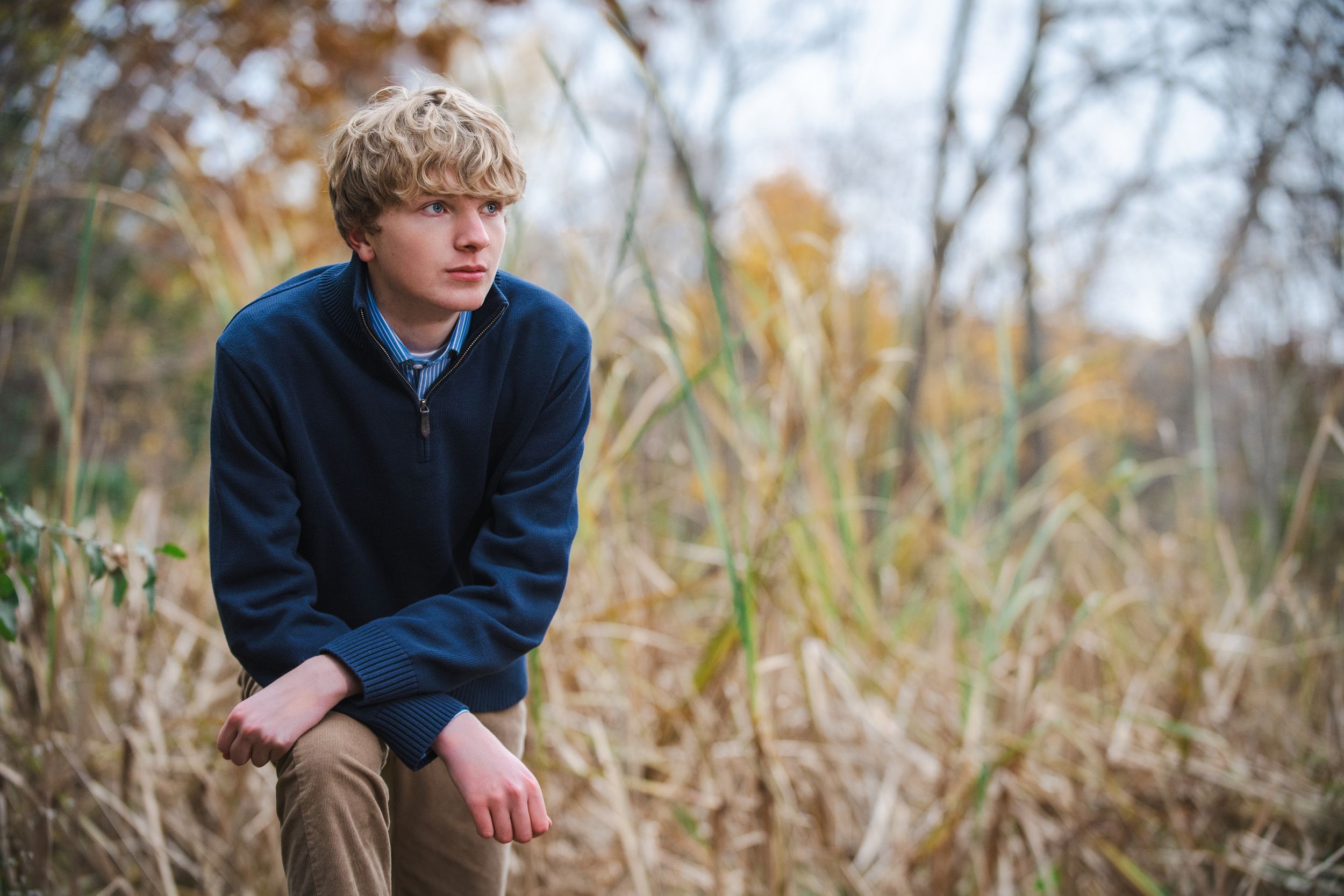 A young blonde teen boy in a navy sweater sits pensively outdoors among tall golden grasses in autumn.