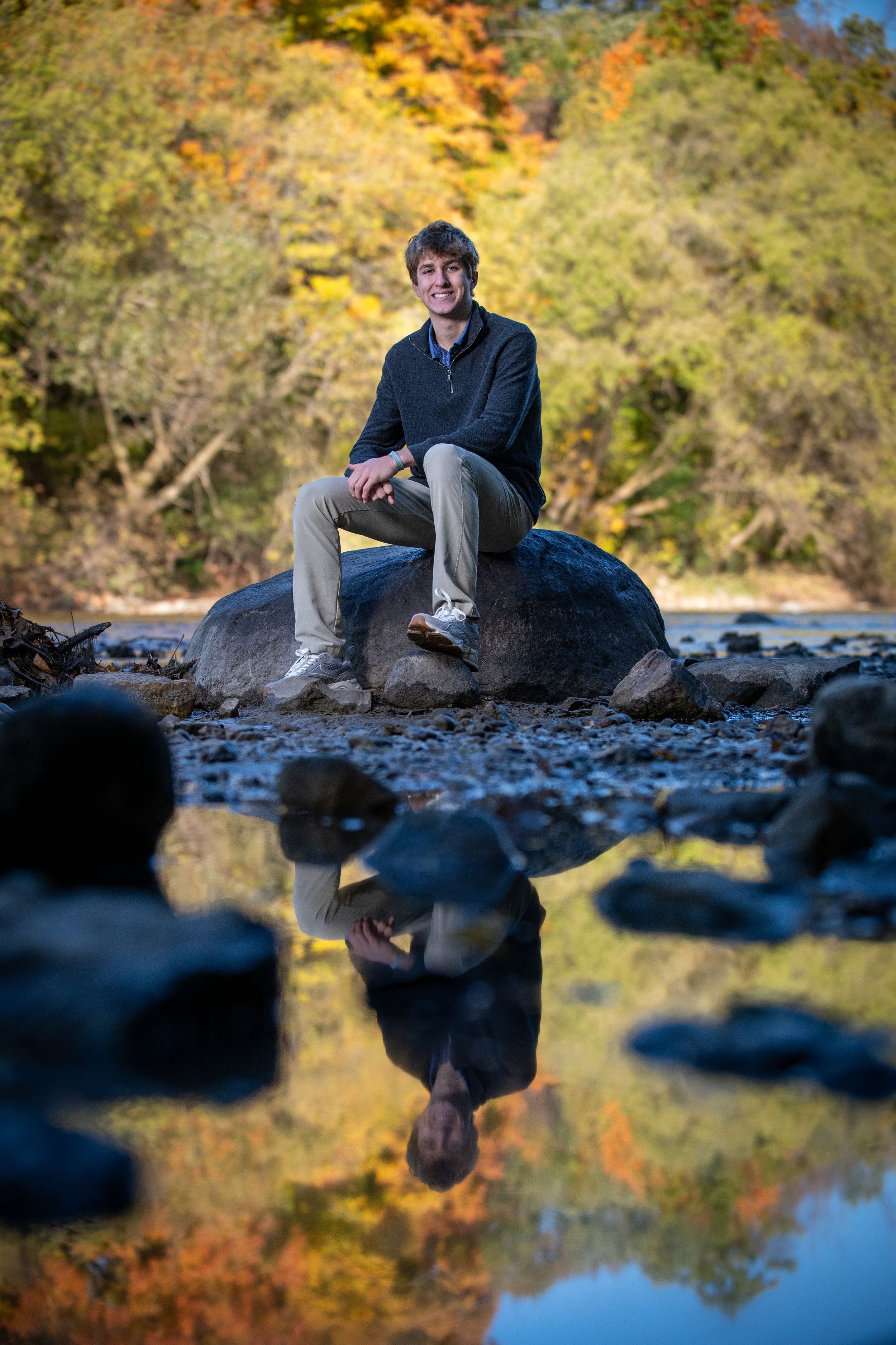 Man sitting on rock by reflective stream with colorful autumn foliage backdrop in nature portrait.